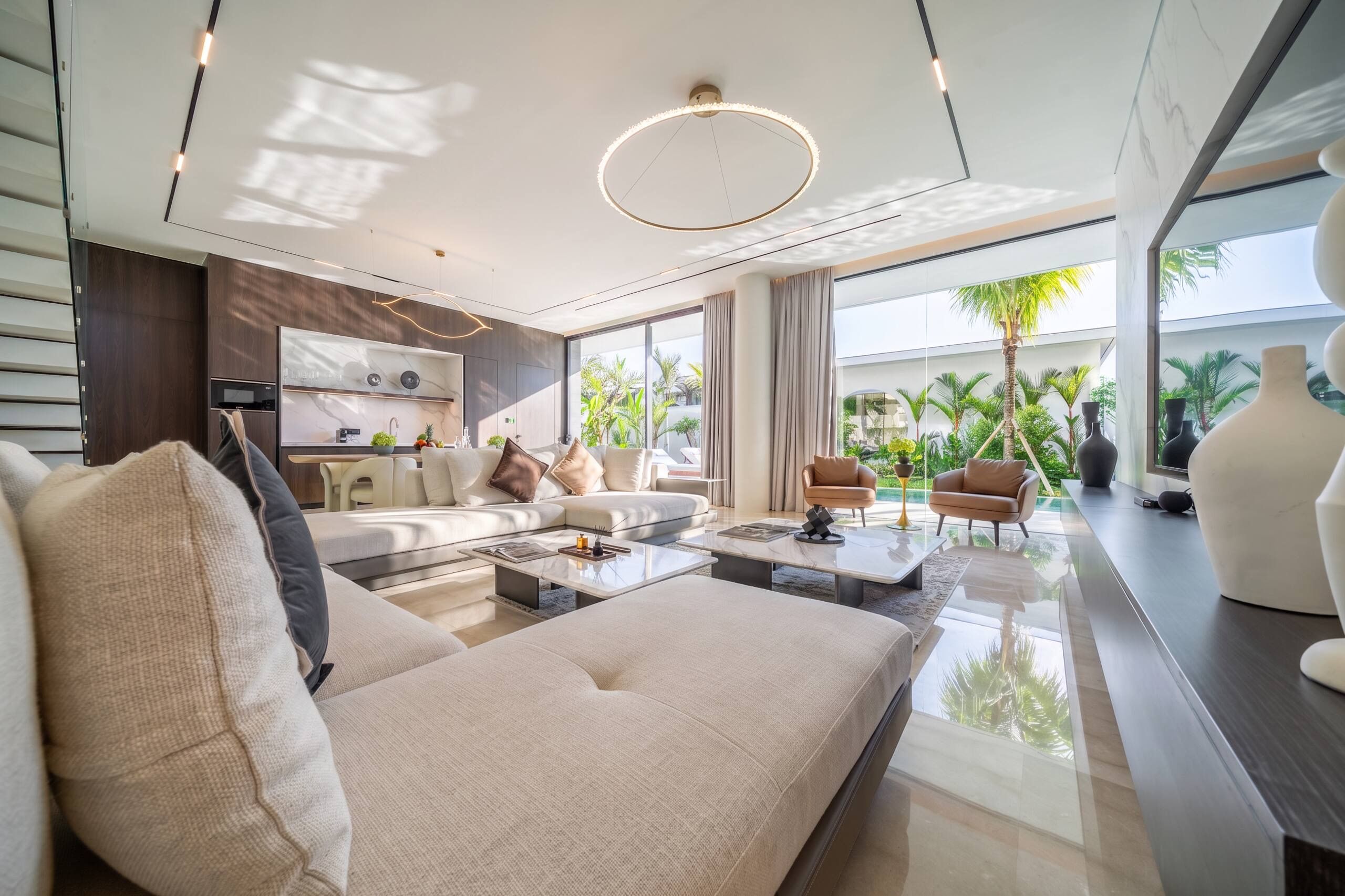 A modern living room with a sofa, coffee table, and chairs, a kitchen area with a dining table, and large windows offering a view of greenery.
