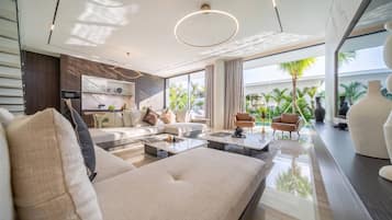 A modern living room with a sofa, coffee table, and chairs, a kitchen area with a dining table, and large windows offering a view of greenery.