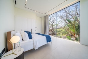 Un dormitorio moderno con un ventanal grande, una cama con ropa de cama blanca y detalles azules, una mesita de noche con una lámpara, y vista a zonas verdes al exterior.