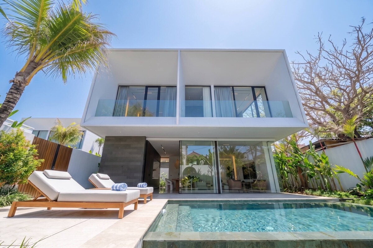 Una casa moderna de dos pisos con piscina, sillas de descanso y plantas tropicales.