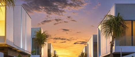 A pathway flanked by modern buildings and lush greenery, leading towards a sunset.
