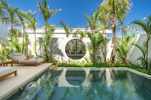 Un espace aménagé au bord de la piscine, avec des chaises longues, une fenêtre circulaire et des plantes tropicales.