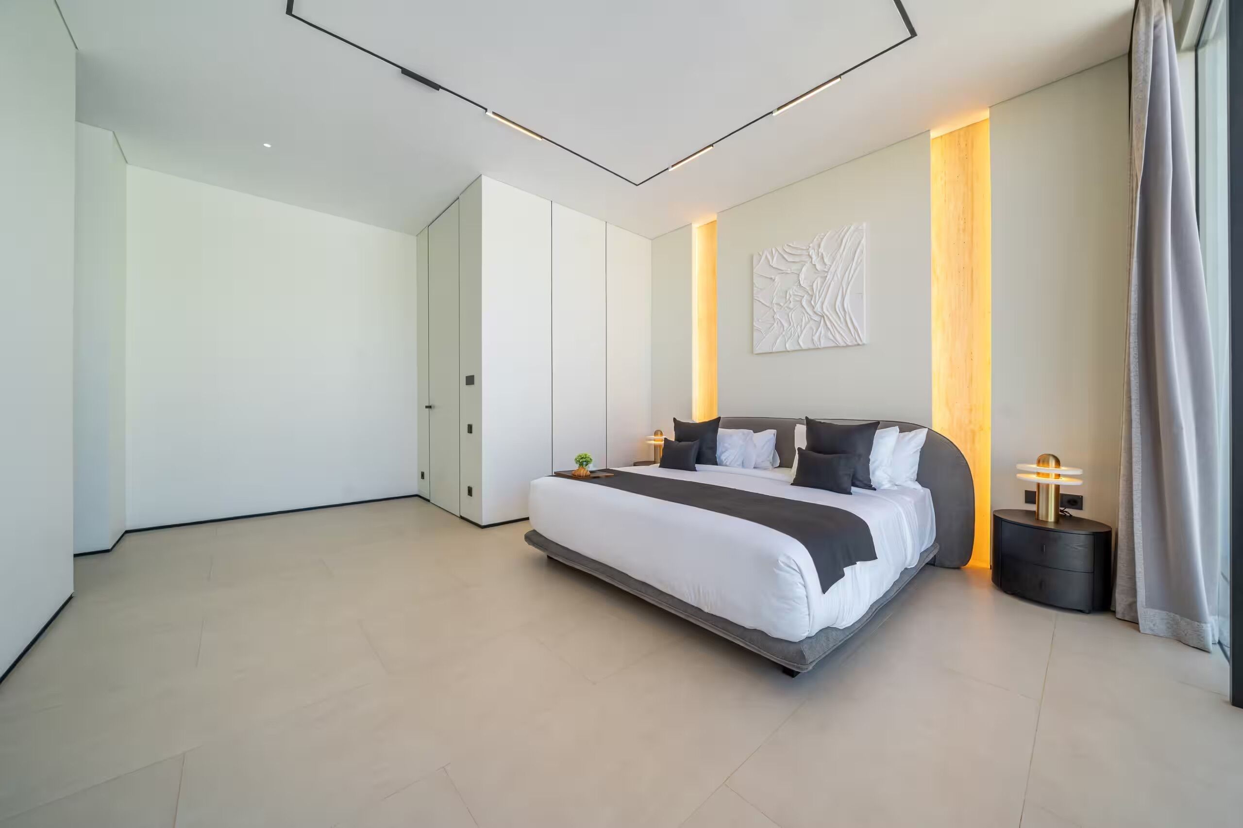 A modern bedroom with a large bed, a bedside table, a wall-mounted light, and a door.