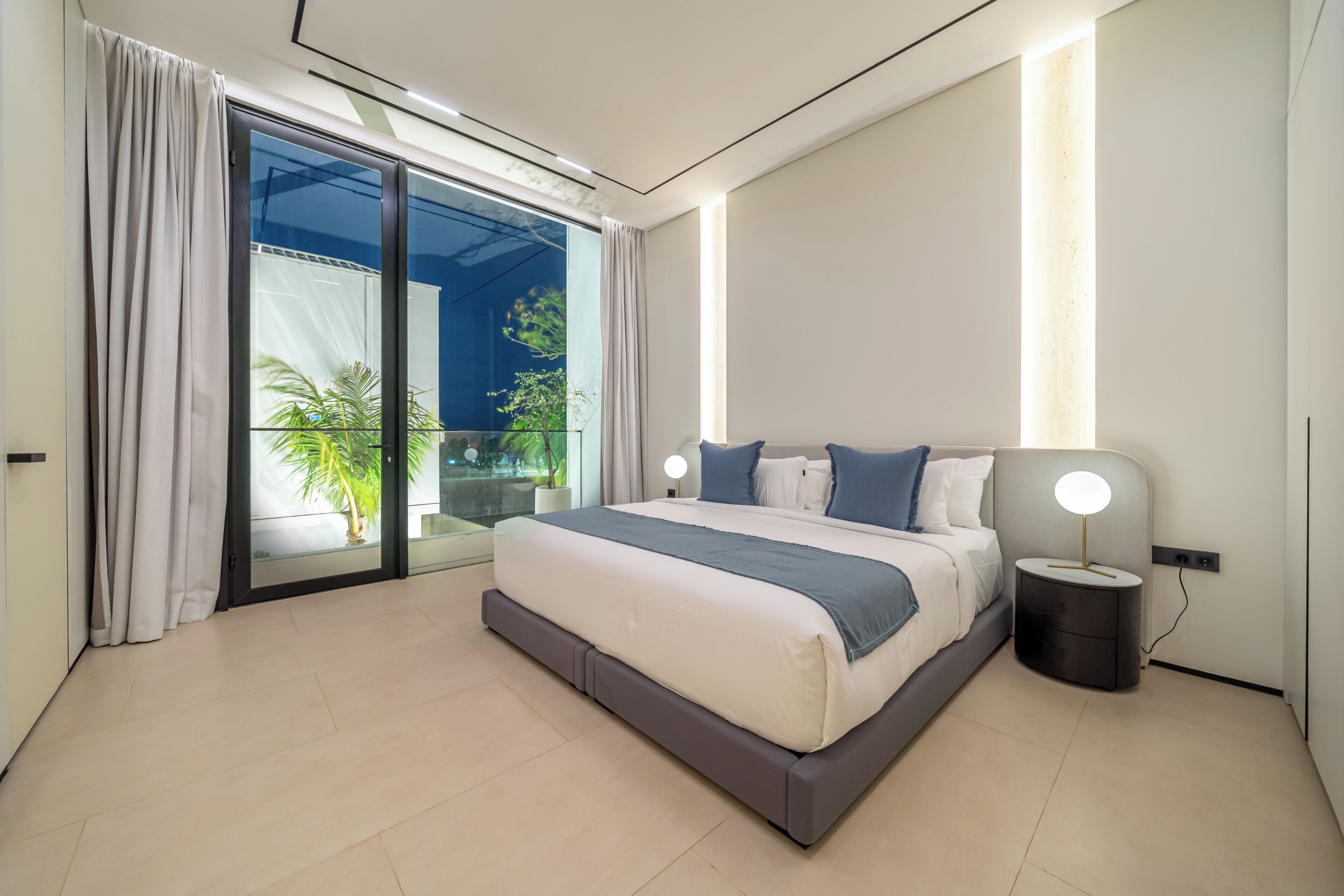Een moderne slaapkamer met een groot bed, een nachtkastje, een lamp en uitzicht op groen via een glazen deur.