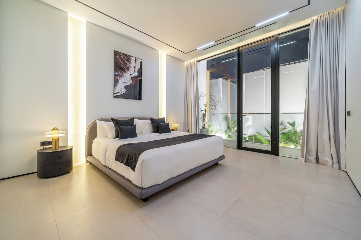 Une chambre moderne avec un grand lit, une table de chevet, une œuvre d’art accrochée au mur et une porte-fenêtre coulissante donnant sur un balcon.