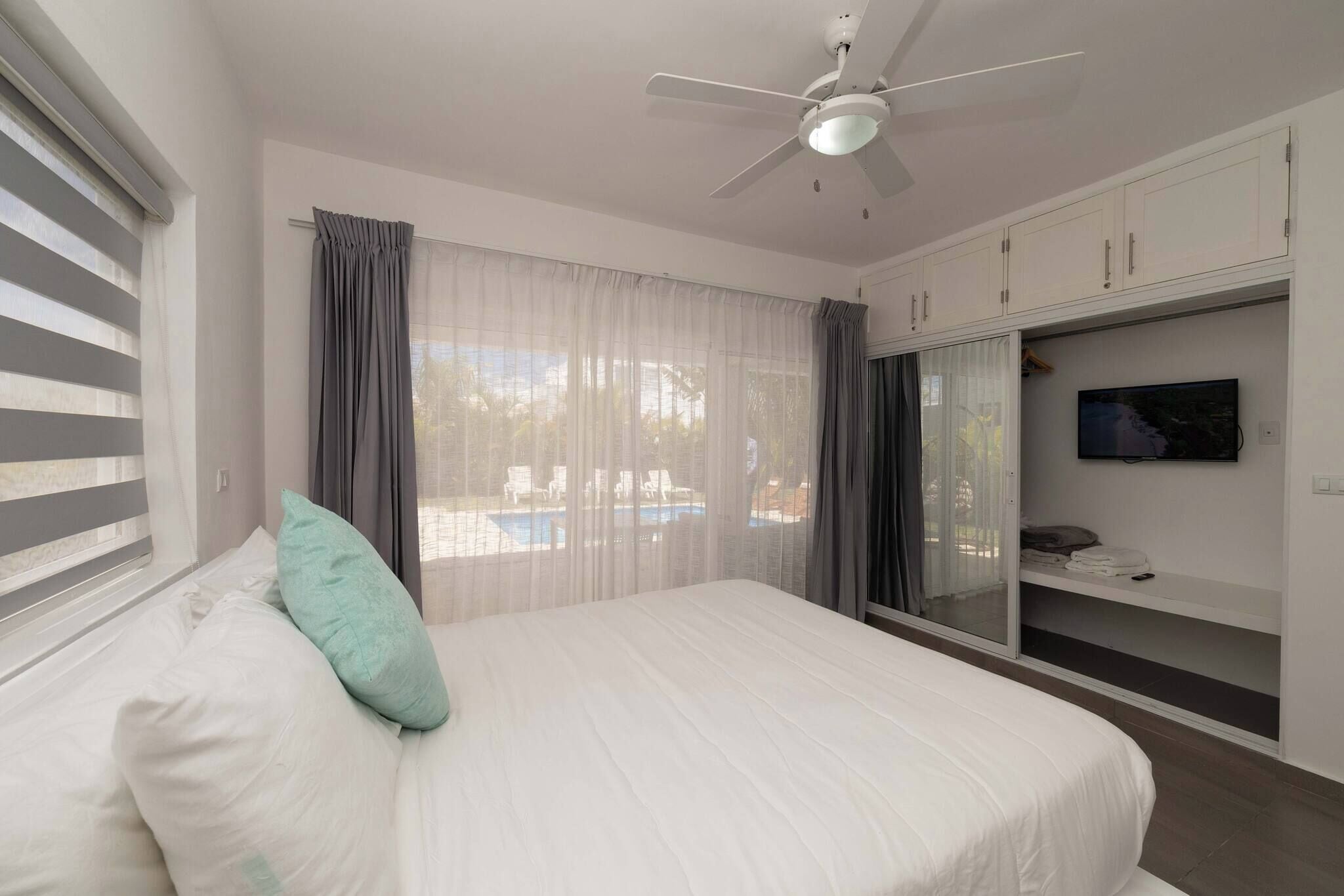 Une chambre avec un lit, un ventilateur de plafond, un téléviseur posé sur un meuble et une vue sur une piscine à l’extérieur.