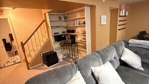 A living room with a grey sofa, a staircase leading to a kitchen, and a wall-mounted shelf with decorative items.