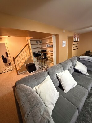 A living room with a grey sofa, a staircase leading to a kitchen, and a wall-mounted shelf with decorative items.