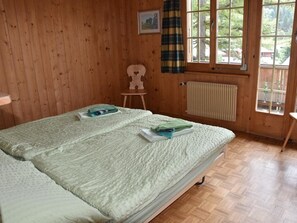 Ein Zimmer mit zwei Betten, einem Heizkörper, einem Fenster mit Vorhängen und einem kleinen Stuhl.