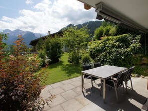 Eine Terrasse im Freien mit Tisch und Stühlen, umgeben von einem gepflegten Garten und Blick auf die Berge.
