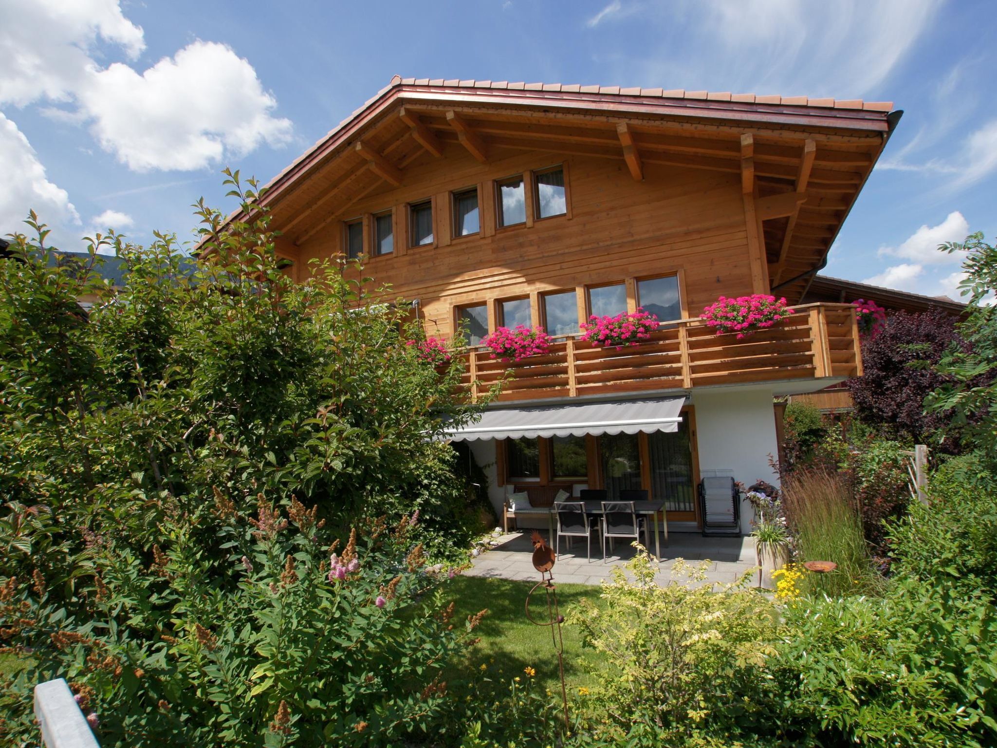 A wooden chalet with a balcony, a covered patio, and a garden with various plants and flowers.
