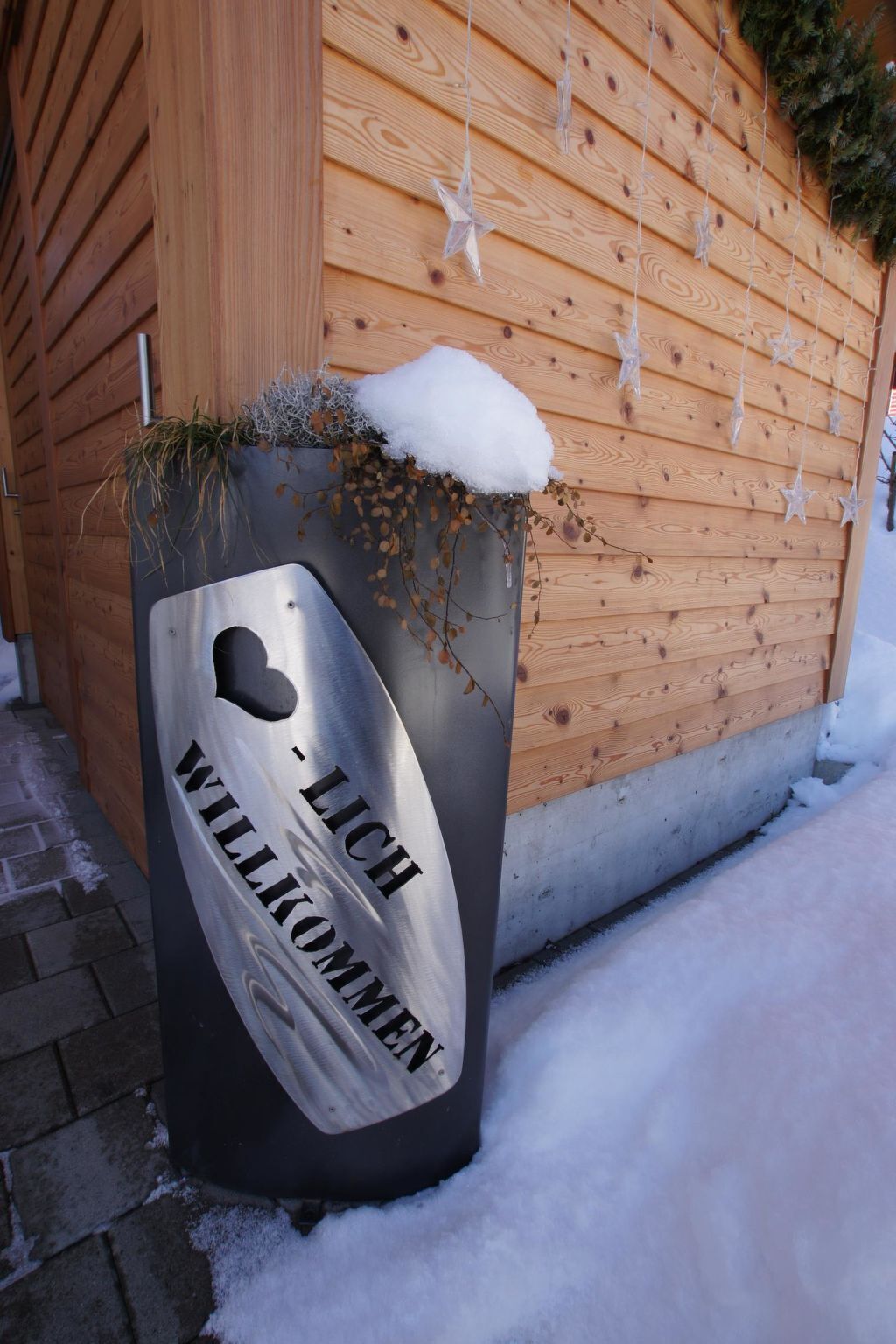 A metal welcome sign with a heart-shaped cutout, placed against a wooden wall with snow on the ground.
