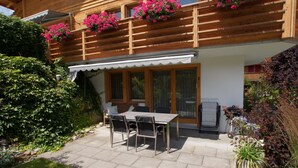 A wooden building with a balcony, a covered patio with a table and chairs, and a garden with various plants. - Sina Appartement/Fewo, Dusche oder Bad, WC, 1 Schlafraum 2-Bett-Wohnung by Interhome (Lenk)