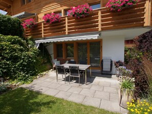 Ein Holzbau mit Balkon, einer überdachten Veranda mit Tisch und Stühlen sowie ein Garten mit verschiedenen Pflanzen.