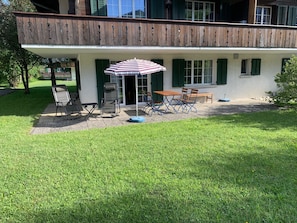 Ein Haus mit einer überdachten Terrasse, ausgestattet mit Tisch und Stühlen, einem gestreiften Sonnenschirm und einem kleinen Garten.