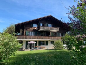 Ein zweigeschossiges Holzhaus mit Balkon, einer überdachten Terrasse mit Stühlen und Sonnenschirm sowie einem Rasenstück.