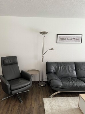 Ein Wohnzimmer mit einem schwarzen Ledersessel, einer schwarzen Ledersofa, einer Stehlampe und einem kleinen runden Tisch. An der Wand hängt ein gerahmtes Schild mit der Aufschrift 'Home sweet Home'.