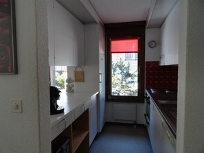 Eine kleine Küche mit weißen Schränken, einer Kaffeemaschine, einem Fenster mit rotem Rollo und einer Uhr an der Wand.