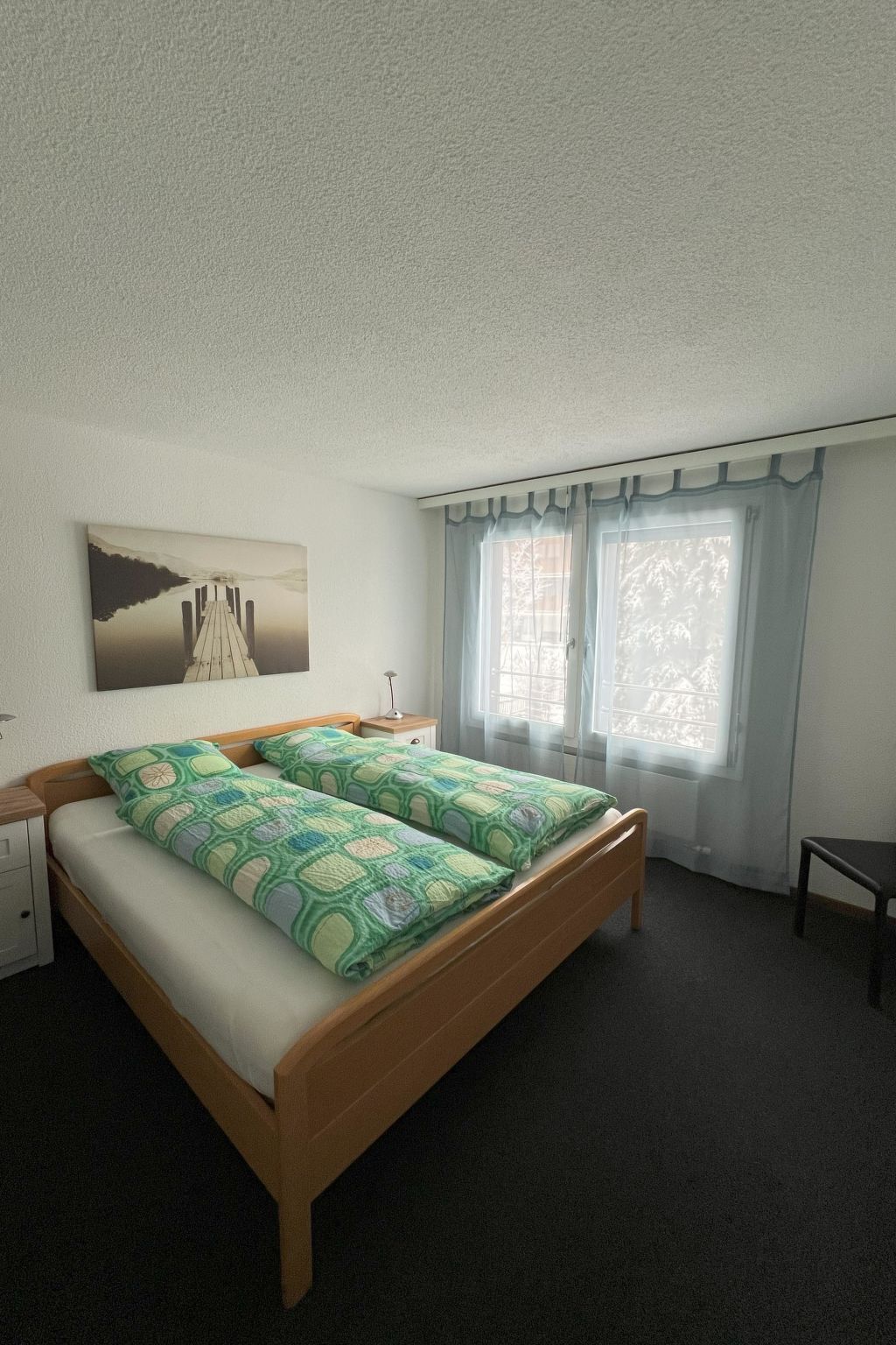 Un dormitorio con cama, mesita de noche, lámpara y ventana con cortinas.