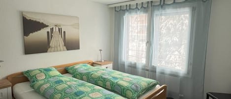 Un dormitorio con cama, mesita de noche, lámpara y ventana con cortinas.