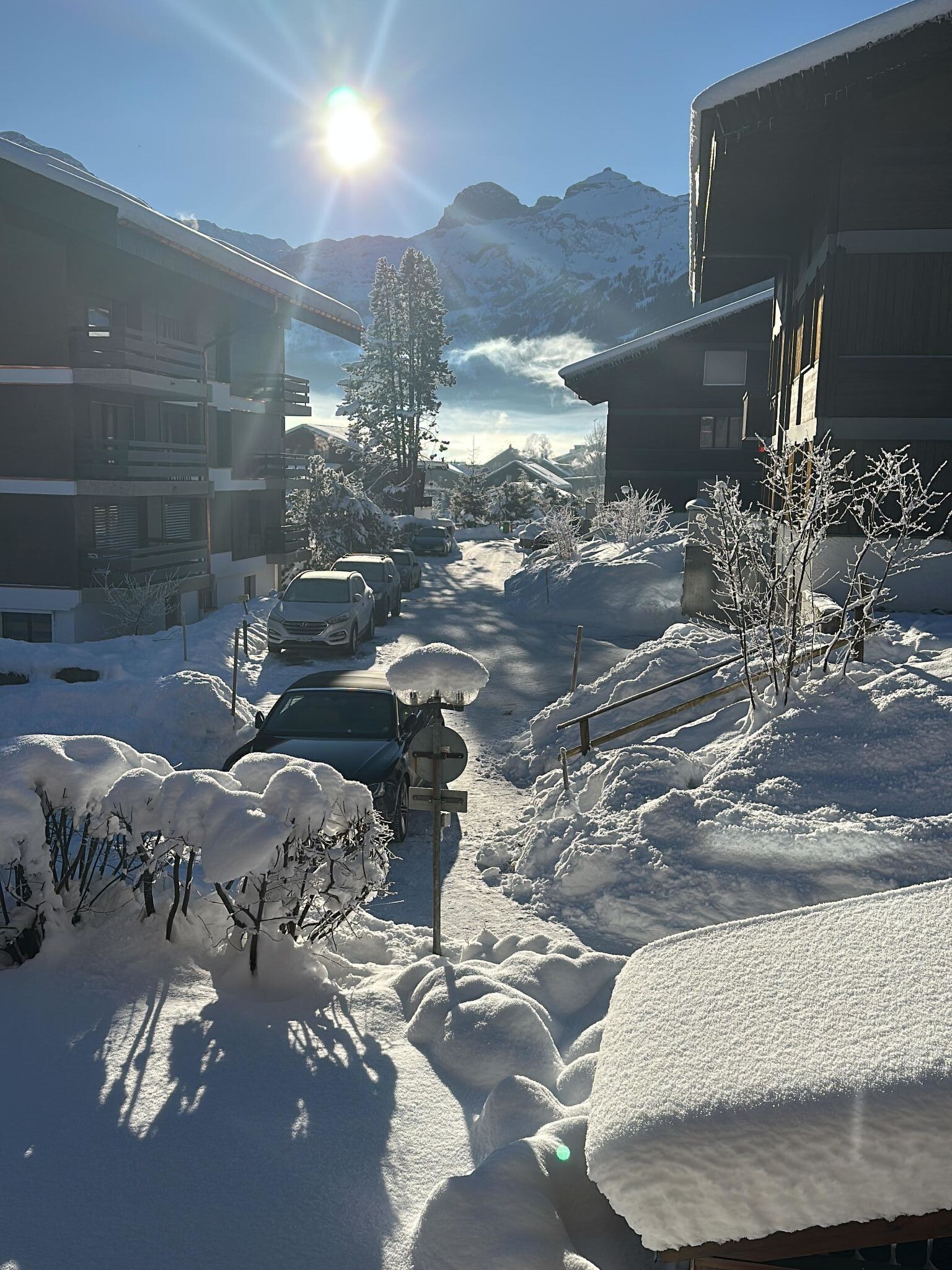 A snow-covered street with buildings, parked cars, and a mountain in the background.