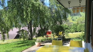 An outdoor patio with a yellow table and red chairs, surrounded by greenery and a view of a village. - Am Brüggli # 1 Appartement/Fewo, Dusche, WC, Erdgeschoß/Parterre 2-Bett-Wohnung by Interhome (Lenk)