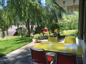 Eine Terrasse im Freien mit einem gelben Tisch und roten Stühlen, umgeben von Grünpflanzen und Blick auf ein Dorf.