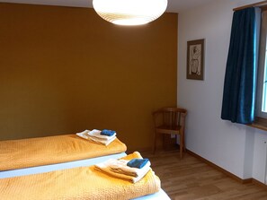 Ein Zimmer mit zwei Betten, einem Sessel, einem Fenster mit Vorhängen und einem Wandbild.