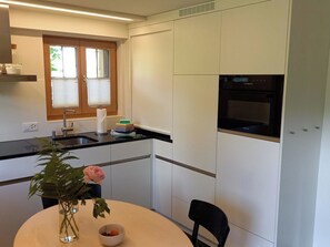 A compact kitchen with a built-in oven, a window with blinds, and a dining area with a table and chairs. - Am Brüggli # 1 Appartement/Fewo, Dusche, WC, Erdgeschoß/Parterre 2-Bett-Wohnung by Interhome (Lenk)