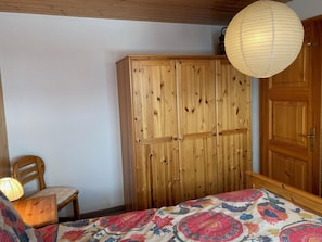 Ein Schlafzimmer mit einem Holzschrank, einem Bett mit bunter Bettdecke, einem Holzstuhl und einer hängenden Lampenleuchte.
