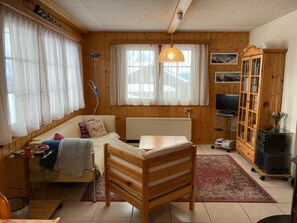 A cozy living room with a wooden floor, a sofa, a wooden chair, a television, a bookshelf, and a window with curtains. - Chalet Pulsatilla Appartement/Fewo, Dusche, WC Pulsatilla by Interhome (Rosswald)