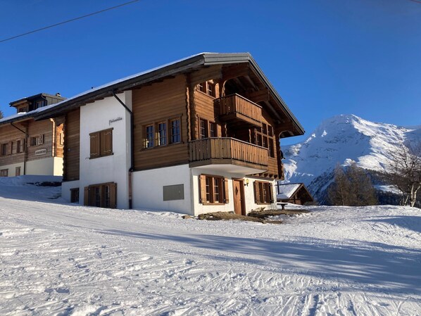 A wooden chalet with a balcony, situated on a snowy slope with a mountain in the background.