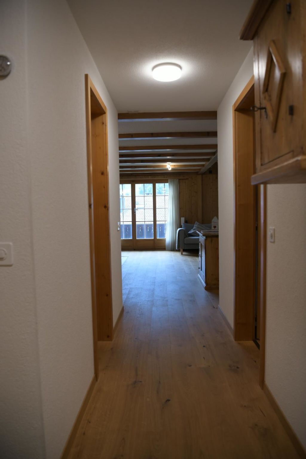 Een hal met houten vloer, een plafondlamp en uitzicht op een keuken met een raam.