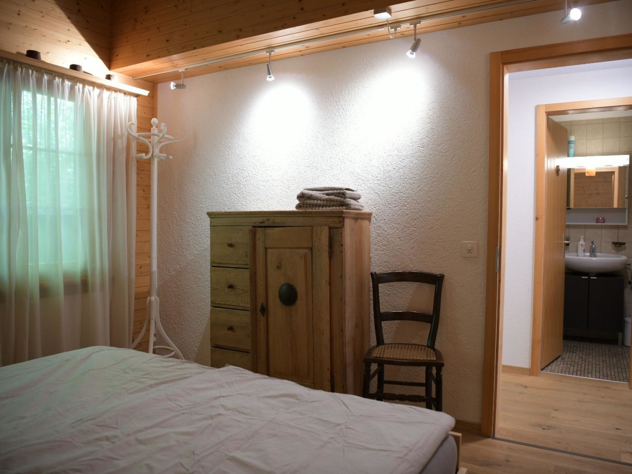 A bedroom with a bed, a wooden wardrobe, a chair, and a bathroom visible through an open door.