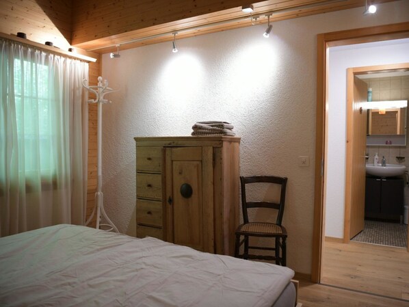 Un dormitorio con cama, un armario de madera, una silla y un baño visible a través de una puerta abierta.