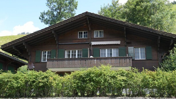 En traditionell stuga i trä med en balkong, belägen i ett frodigt, grönt landskap.