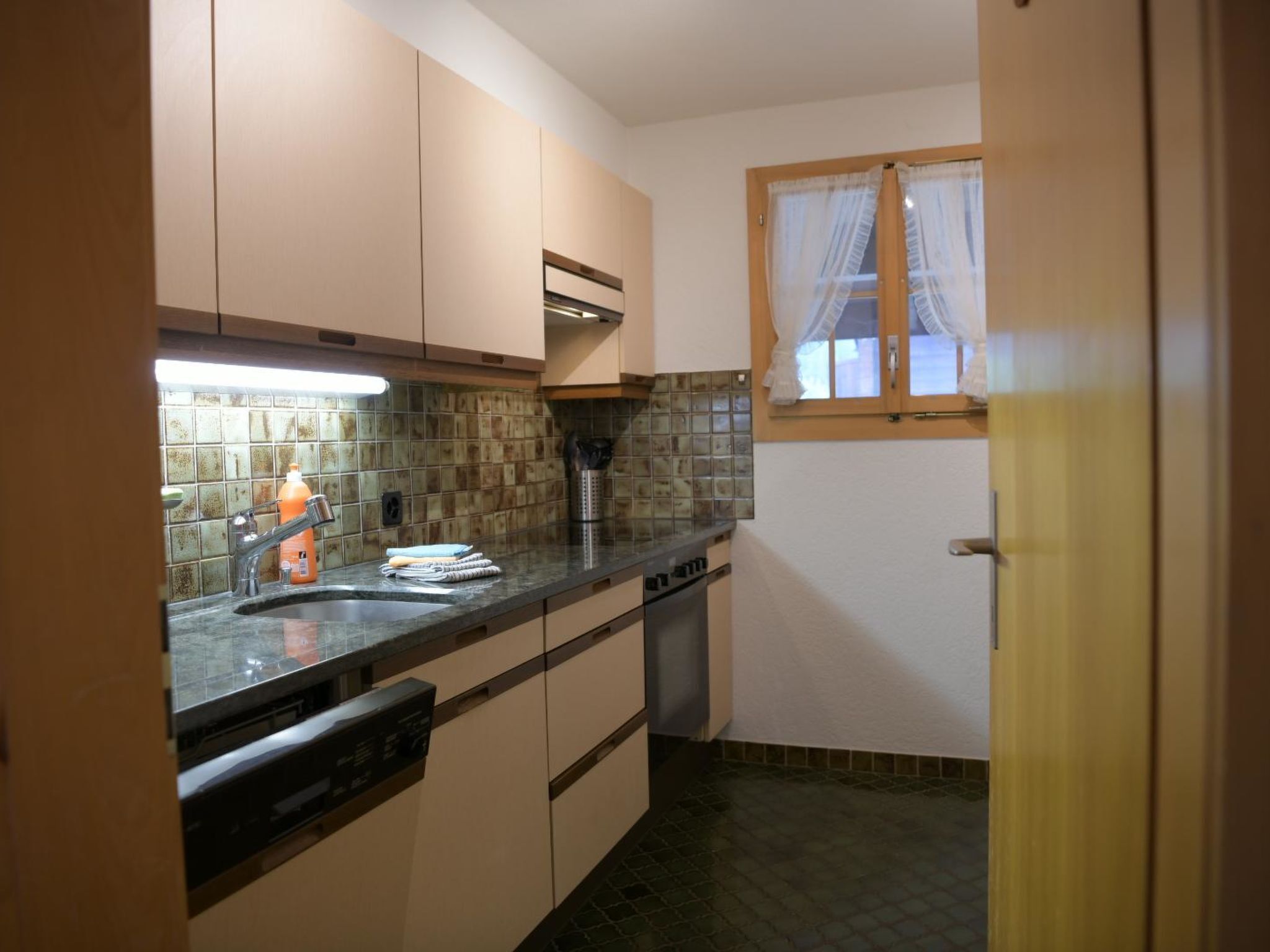 A compact kitchen with a dishwasher, a sink, and a window with curtains.