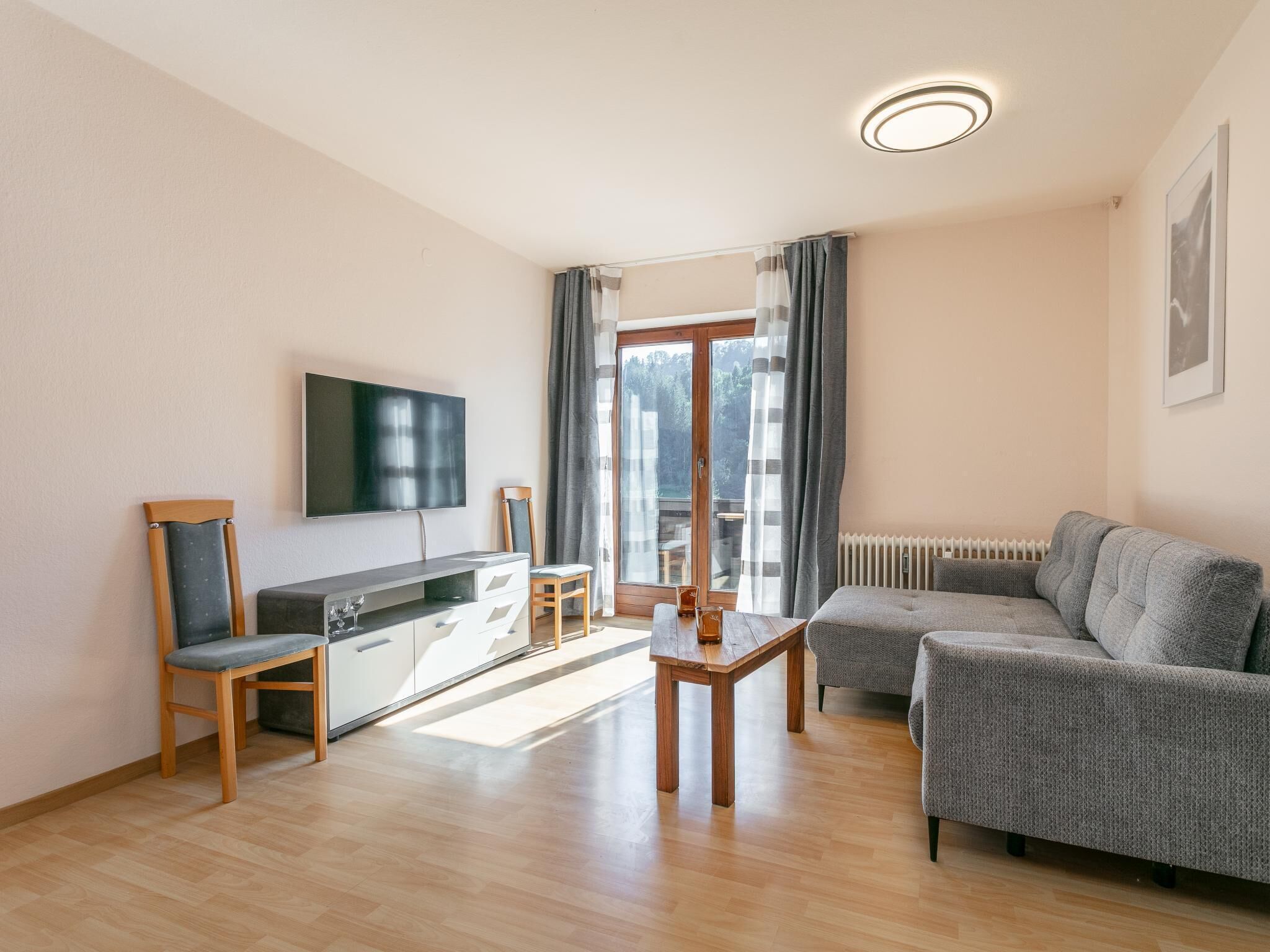 Uma sala de estar moderna com sofá, mesa de jantar, TV fixada na parede e vista para o exterior através de uma grande janela.