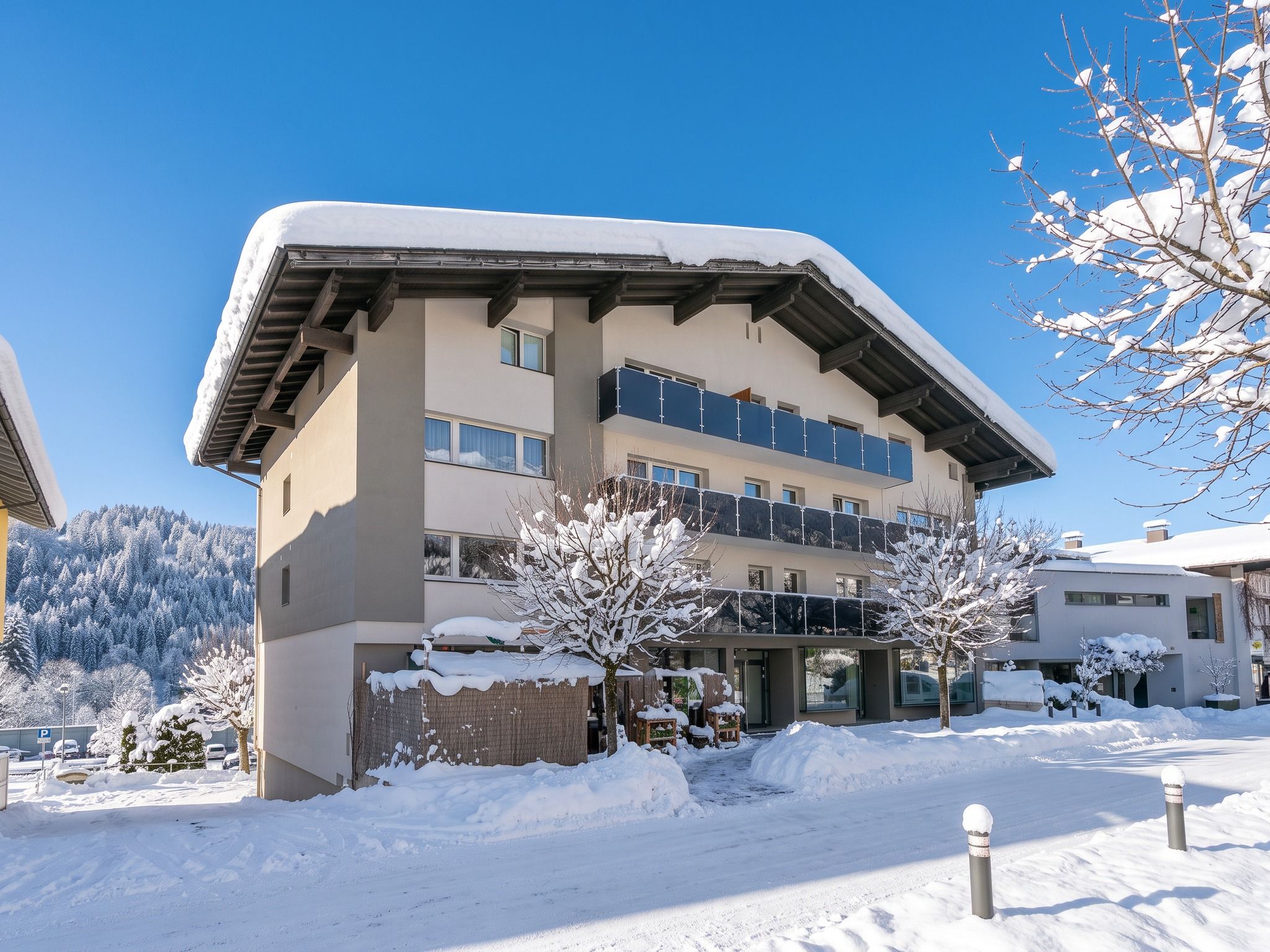 Een hotel met meerdere verdiepingen, met een met sneeuw bedekte sneeuwkap en balkons, omringd door besneeuwde bomen en een bergachtige achtergrond.