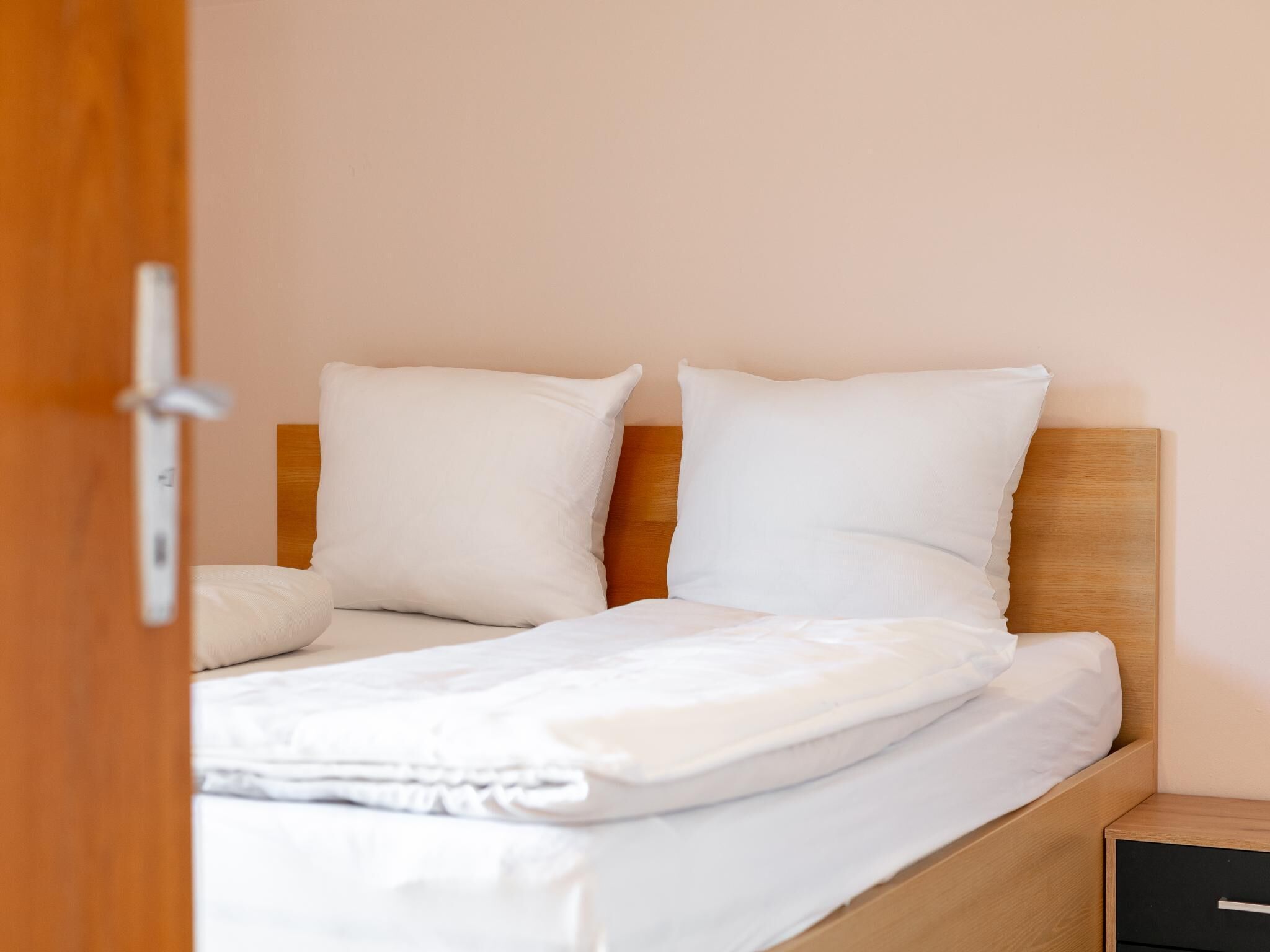A neatly made bed with white bedding and pillows, a wooden headboard, and a small bedside table with a drawer.