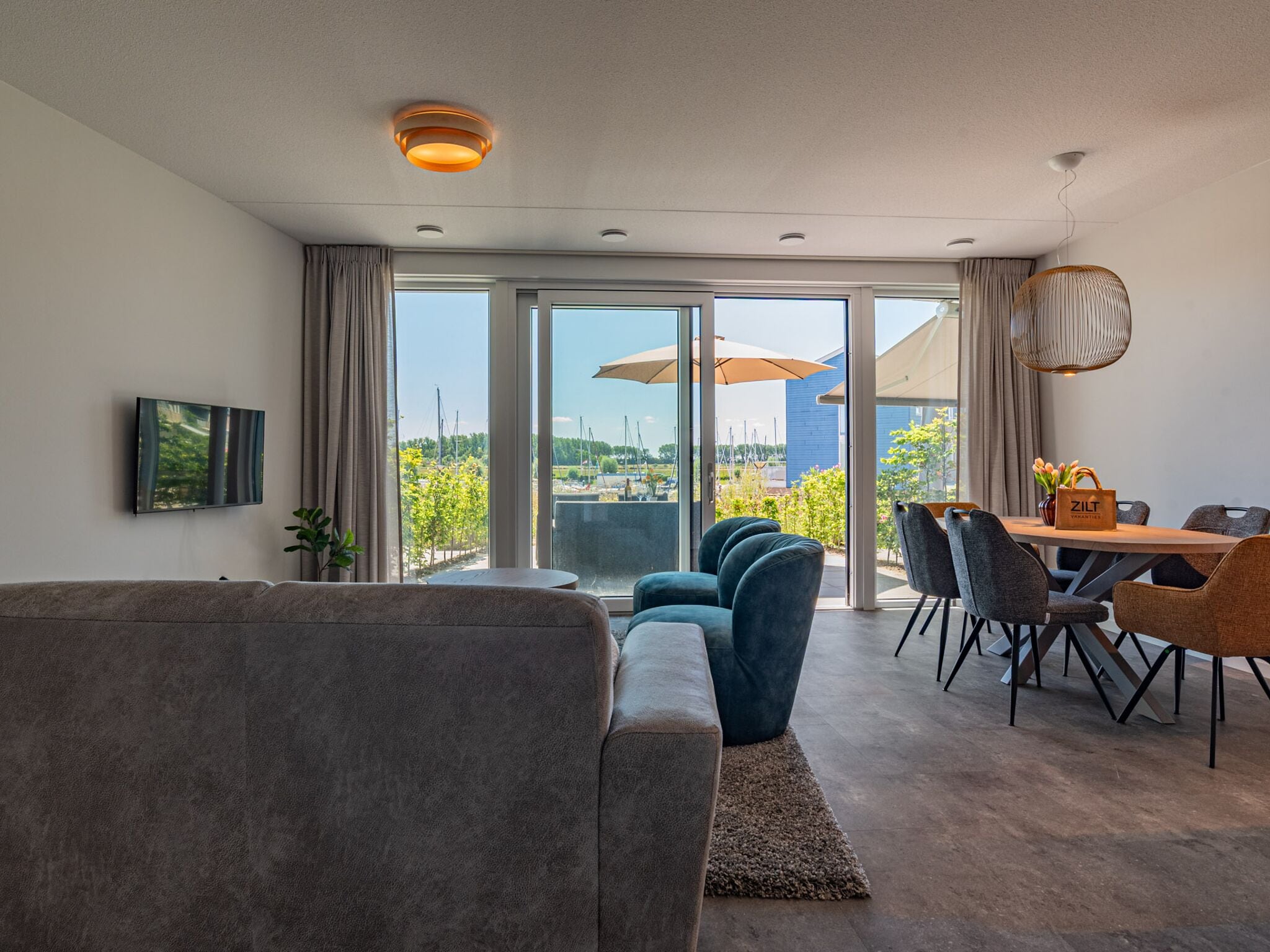 A modern living room with a sofa, a dining table with chairs, and a view of a marina through large glass doors.