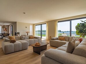 A spacious living room with two sofas, a coffee table, and a large window offering a view of a lake. - Luxury villa by the Veerse Meer (Kamperland)