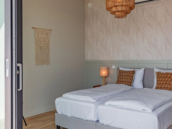 Un letto matrimoniale con lenzuola bianche, una lampada da comodino, cuscini decorati e un arazzo in macramè in una stanza con parete strutturata e lampadario intrecciato.