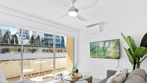Ein modernes Wohnzimmer mit einem Glastisch, grauen Sofas, einem Deckenventilator, einem an der Wand montierten Fernseher und einem Balkon mit Blick auf Gebäude und Bäume.