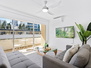 A modern living room with a glass coffee table, grey sofas, a ceiling fan, a TV mounted on the wall, and a balcony with a view of buildings and trees. - Beachfront Escape Broadbeach (Broadbeach)