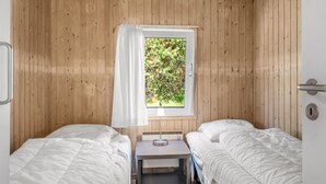 Ett litet, enkelt rum med två sängar, ett fönster med utsikt över grönområden, ett nattduksbord med en lampa och en dörr med ett handtag.