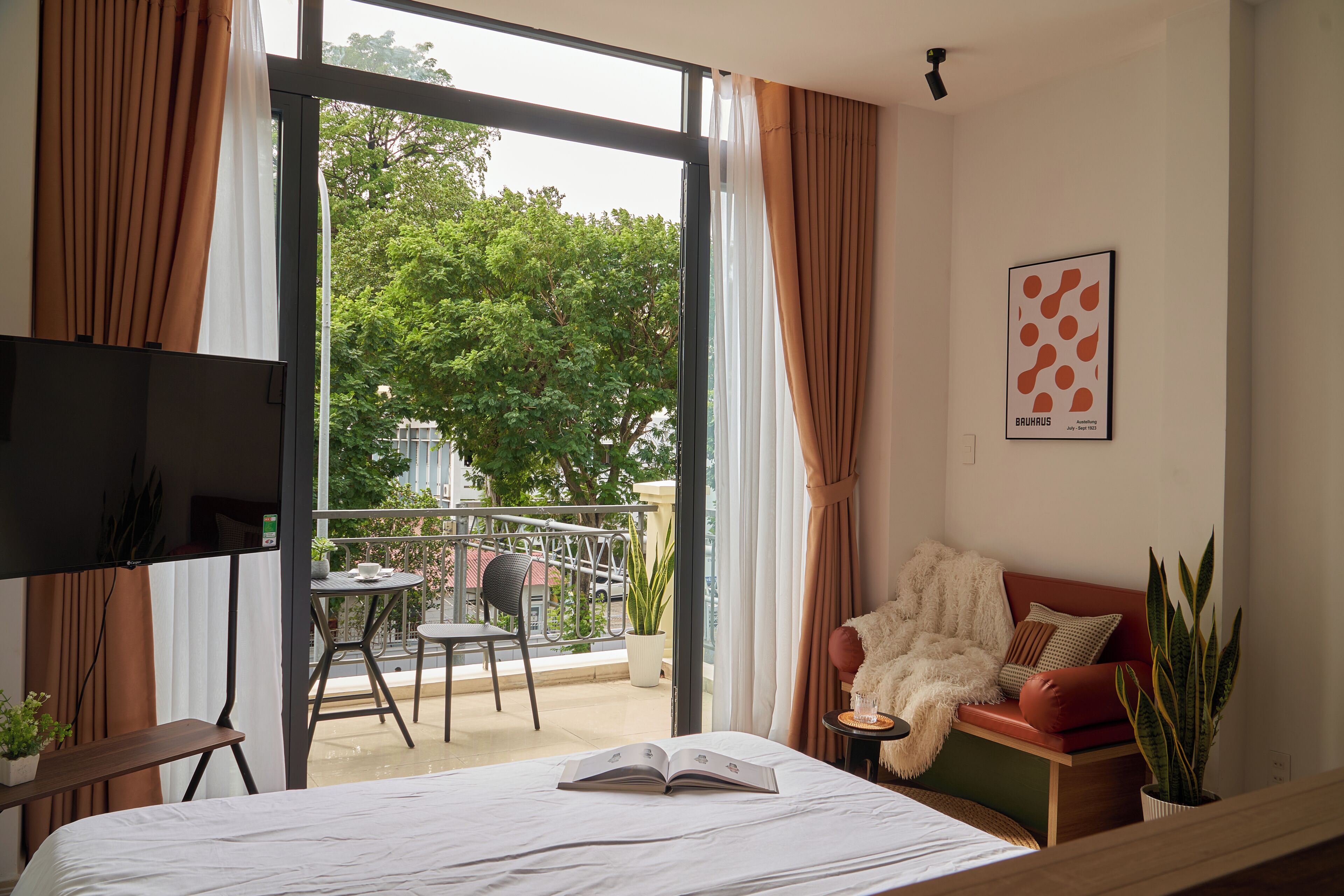 A hotel room with a bed, a TV, a balcony with a chair and table, and a view of trees and buildings outside.