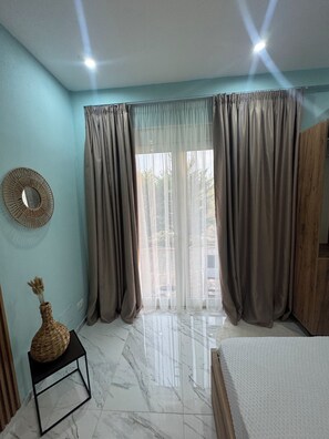 A bedroom with a bed, a small table, a mirror, and a view through a window with curtains. - Calm and Green Retreat (Velipojë)