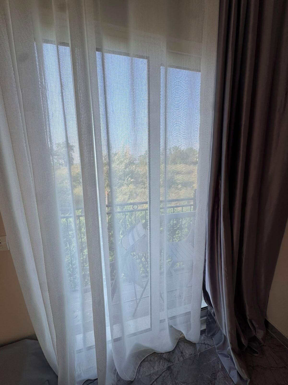 Une pièce avec vue sur l’extérieur à travers de fines rideaux, un balcon et un plancher foncé.