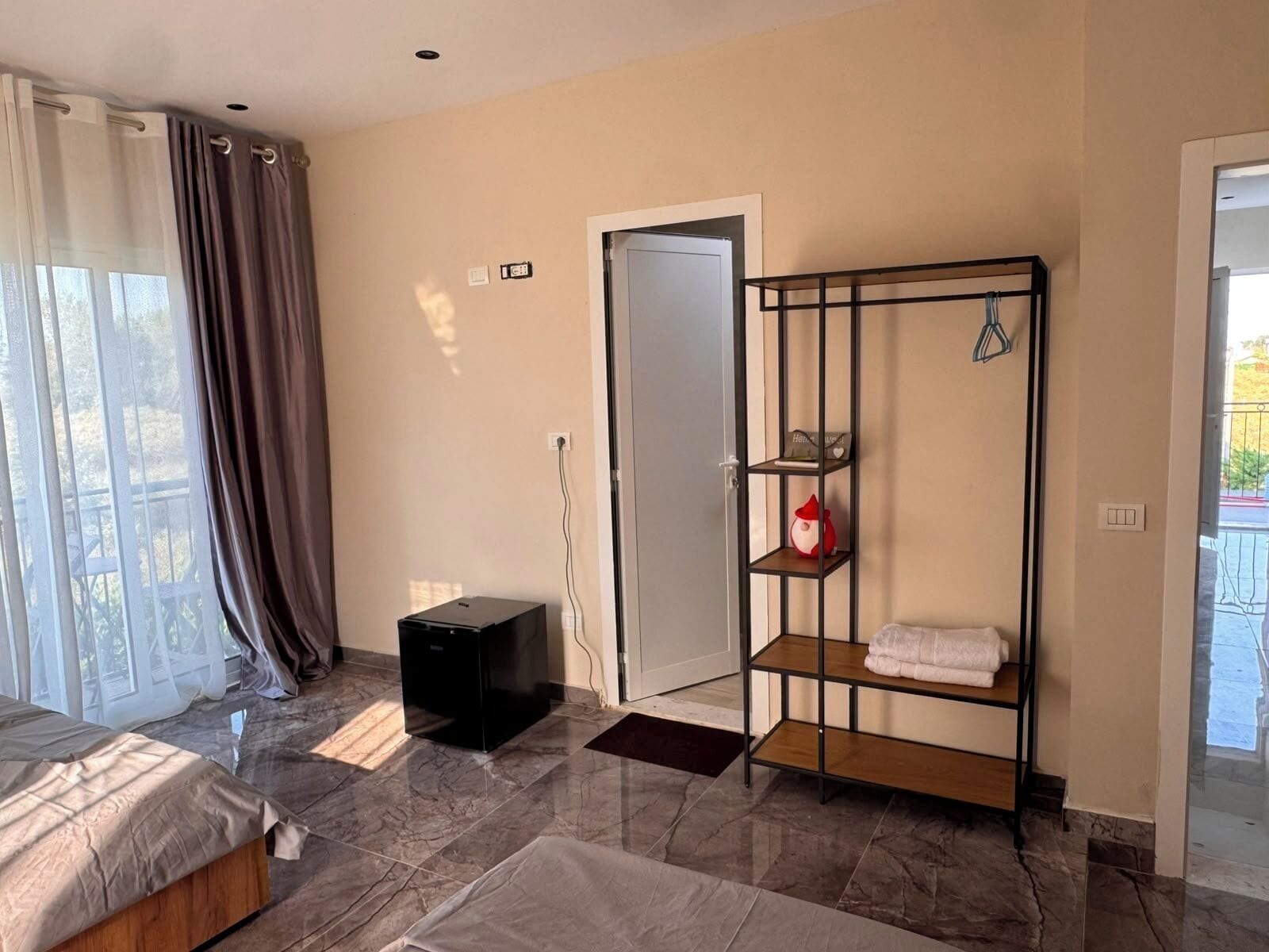 Habitación de hotel con cama, armario, un mueble negro pequeño, estantería y puerta que da a un balcón.